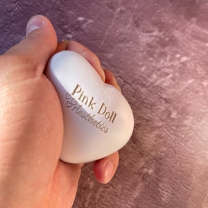 K&ouml;nnte beinhalten: Ein wei&szlig;er herzf&ouml;rmiger Stressball mit dem Aufdruck "Pink Doll Aesthetics".