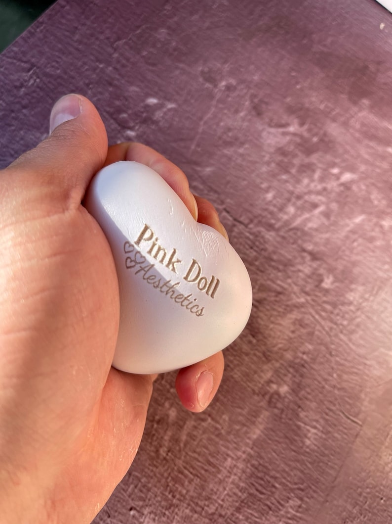 K&ouml;nnte beinhalten: Ein wei&szlig;er herzf&ouml;rmiger Stressball mit dem Aufdruck "Pink Doll Aesthetics".
