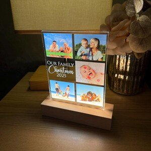 Puede incluir: Un expositor de fotos rectangular con cuatro fotos familiares, montado sobre una base de madera. El texto "OUR FAMILY CHRISTMAS 2023" est&aacute; impreso en la parte delantera. La pantalla est&aacute; iluminada desde abajo, creando un brillo c&aacute;lido.