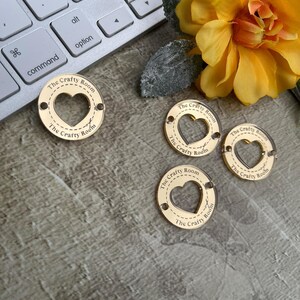 May include: Four gold-coloured wooden heart-shaped tags with the text "The Crafty Room" printed on each. The tags have a cut-out heart shape in the centre.