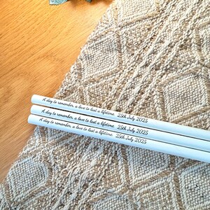 May include: Three white pencils with the inscription "A day to remember, a love to last a lifetime. 25th July 2025". The pencils are placed on a textured, beige and brown woven surface. A yellow flower is in the background.