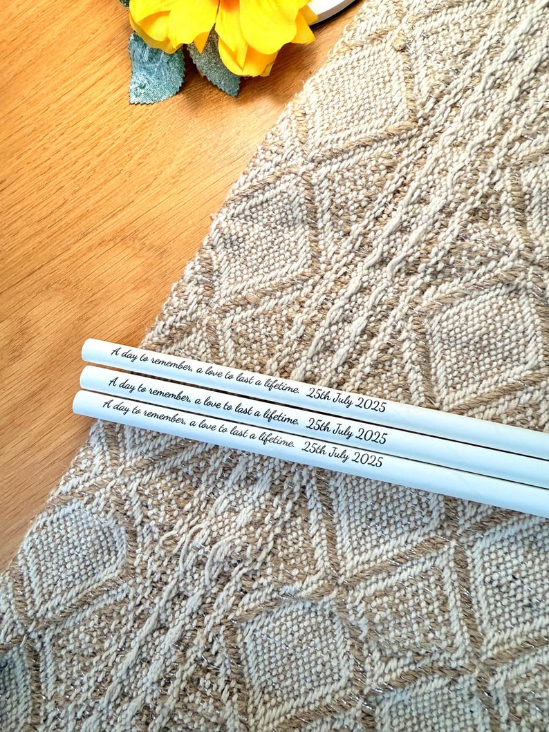 May include: Three white pencils with the inscription "A day to remember, a love to last a lifetime. 25th July 2025". The pencils are placed on a textured, beige and brown woven surface. A yellow flower is in the background.