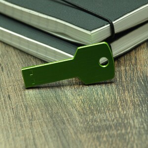 May include: A green metal key-shaped USB drive on a wooden surface. The USB drive is lying on top of two closed black notebooks.