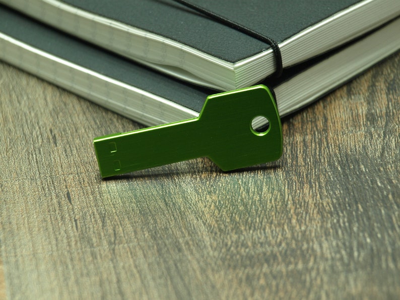 May include: A green metal key-shaped USB drive on a wooden surface. The USB drive is lying on top of two closed black notebooks.