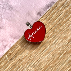 May include: A red heart-shaped charm with a silver ring and the name "Ariana" written in white.