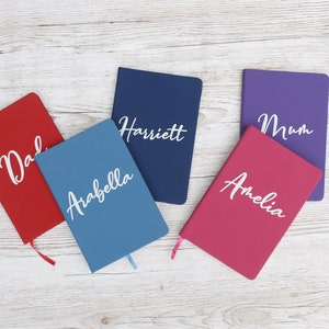 May include: Five personalised notebooks with different names on each. The notebooks are red, blue, purple, pink and navy blue.