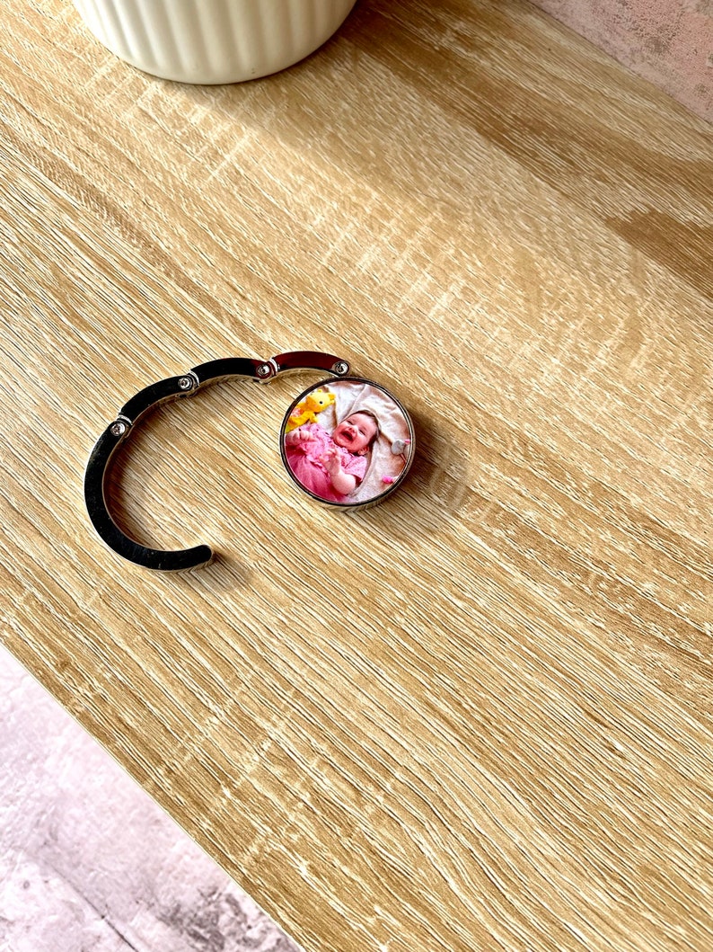May include: A silver metal bag charm with a round photo insert featuring a baby in a pink outfit. The photo insert is attached to a silver metal chain.