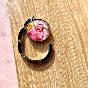 May include: A silver metal keychain with a round photo insert featuring a baby girl in a pink dress. The photo shows the baby smiling and holding a yellow stuffed animal.
