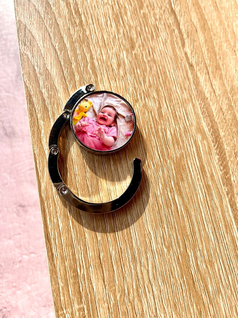 May include: A silver metal keychain with a round photo insert featuring a baby girl in a pink dress. The photo shows the baby smiling and holding a yellow stuffed animal.