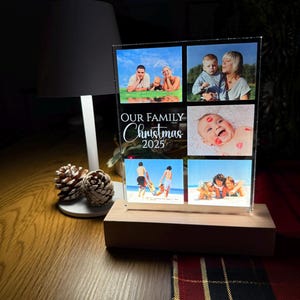 Puede incluir: Una pantalla fotogr&aacute;fica de acr&iacute;lico transparente con seis fotos familiares, colocada sobre una base de madera. El texto "OUR FAMILY Christmas 2025" est&aacute; impreso en la parte delantera. La pantalla est&aacute; iluminada por una luz, creando un brillo c&aacute;lido.