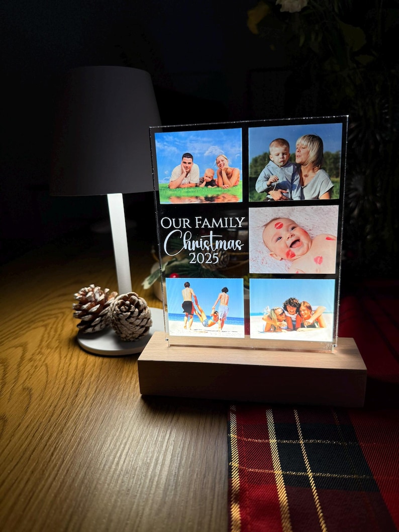 Puede incluir: Una pantalla fotogr&aacute;fica de acr&iacute;lico transparente con seis fotos familiares, colocada sobre una base de madera. El texto "OUR FAMILY Christmas 2025" est&aacute; impreso en la parte delantera. La pantalla est&aacute; iluminada por una luz, creando un brillo c&aacute;lido.