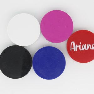May include: Five round phone grips in different colours. The grips are white, black, pink, blue and red. The red grip has the name "Ariana" written in white letters.