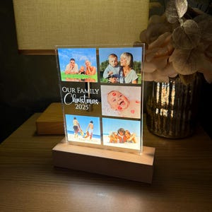 Puede incluir: Un expositor de fotos iluminado con cuatro fotos familiares. El texto "OUR FAMILY Christmas 2025" est&aacute; escrito en blanco. La base es de madera clara y las fotos est&aacute;n iluminadas desde dentro.