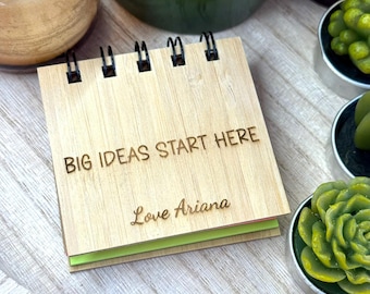 May include: A small wooden notepad with the words "BIG IDEAS START HERE" and "Love Ariana" engraved on the cover. The notepad has a spiral binding and contains green and pink pages. The notepad is on a wooden tray.