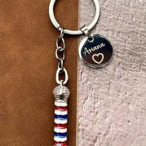 May include: A silver keyring with a barber pole charm and a round tag. The barber pole charm is red, white, and blue. The tag is engraved with the name "Ariana" and a heart symbol. The keyring is on a brown background.