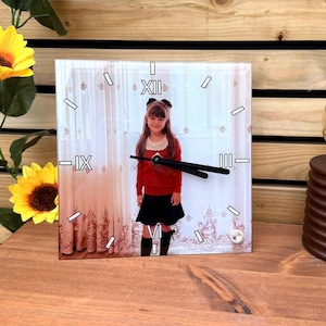 May include: Square clock featuring a photograph of a girl in a red sweater and black skirt. The clock face has Roman numerals and black hands. The background shows a window with curtains.