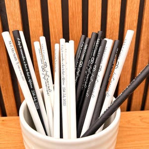 May include: A collection of black and white paper straws in a white ceramic cup. The straws are printed with wedding-themed text, including names, dates, and the phrase "Just Married!" The background is a wooden wall.