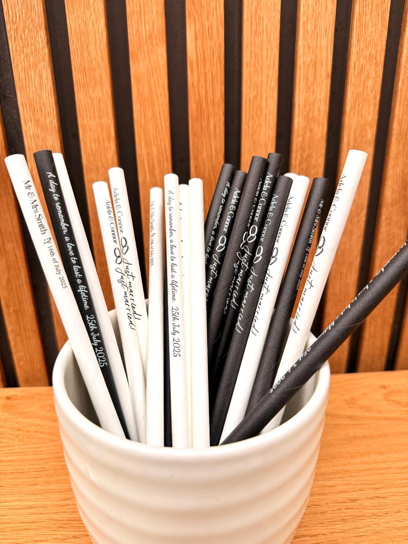 May include: A collection of black and white paper straws in a white ceramic cup. The straws are printed with wedding-themed text, including names, dates, and the phrase "Just Married!" The background is a wooden wall.