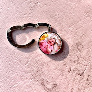 May include: A silver metal keychain with a round photo charm featuring a baby girl smiling in a pink dress. The photo charm is attached to a silver metal hook.