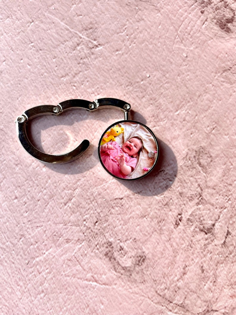 May include: A silver metal keychain with a round photo charm featuring a baby girl smiling in a pink dress. The photo charm is attached to a silver metal hook.