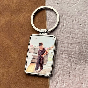 May include: A silver rectangular keyring featuring a photograph of a young girl in a floral dress. The keyring has a silver ring at the top. The background is a brown leather-like surface and a grey textured surface.