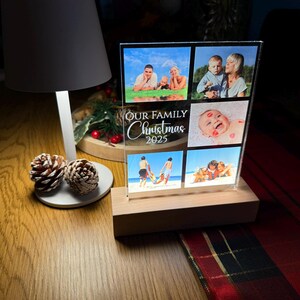 Puede incluir: Una pantalla de fotos de acr&iacute;lico transparente con seis fotos familiares, colocada sobre una base de madera. El texto "OUR FAMILY Christmas 2025" est&aacute; impreso en la parte delantera. La pantalla est&aacute; iluminada por una l&aacute;mpara y pi&ntilde;as.