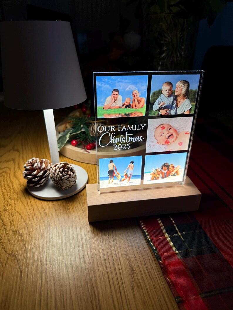 Puede incluir: Una pantalla de fotos de acr&iacute;lico transparente con seis fotos familiares, colocada sobre una base de madera. El texto "OUR FAMILY Christmas 2025" est&aacute; impreso en la parte delantera. La pantalla est&aacute; iluminada por una l&aacute;mpara y pi&ntilde;as.