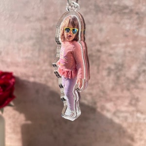 May include: A clear acrylic keychain with a photo of a young girl wearing sunglasses and a pink shirt. The keychain is shaped like a person.