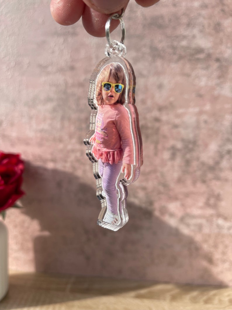 May include: A clear acrylic keychain with a photo of a young girl wearing sunglasses and a pink shirt. The keychain is shaped like a person.