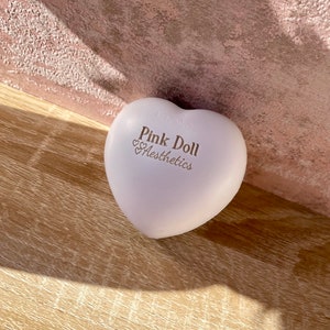 K&ouml;nnte beinhalten: Ein hellrosa, herzf&ouml;rmiges, weiches Spielzeug mit dem aufgedruckten Text "Pink Doll Aesthethics".