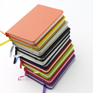 May include: A stack of colourful notebooks with ribbon bookmarks. The top notebook is orange, and the stack includes yellow, silver, black, navy blue, red, green, pink, and purple notebooks.