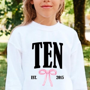 May include: A white long-sleeve sweatshirt featuring the black text TEN centered on the chest. Below the text is a small pink bow graphic flanked by the black text EST. on the left and 2015 on the right. The garment has a crew neckline.