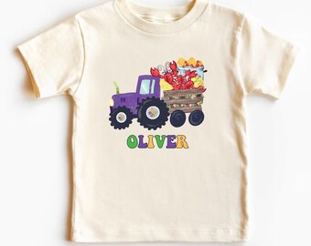 Puede incluir: Camiseta color crema con un tractor morado que tira de un vagón lleno de cangrejos rojos y maíz amarillo. El nombre "OLIVER" está impreso en letras coloridas debajo. Diseño ilustrativo y divertido.