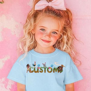 A young girl with blonde hair wearing a light blue t-shirt with a cartoon character design and a pink bow in her hair.