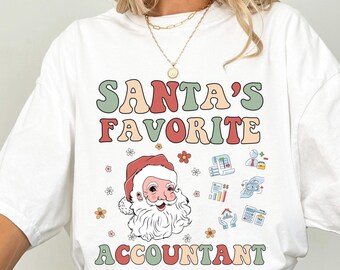 May include: A white short-sleeve t-shirt featuring the text SANTA'S FAVORITE ACCOUNTANT in retro-style lettering. The design includes a graphic of Santa Claus surrounded by small flower icons and various accounting-themed symbols and charts.
