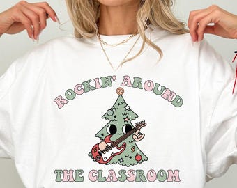 May include: A white t-shirt featuring a graphic of a cartoon Christmas tree playing a red electric guitar. The text "ROCKIN' AROUND THE CLASSROOM" is printed in pink and green letters above and below the tree illustration.