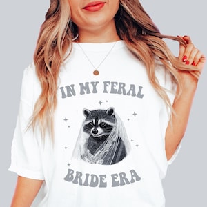 May include: White t-shirt with a graphic of a raccoon wearing a bridal veil and the text "In My Feral Bride Era".
