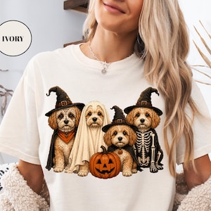 May include: An ivory t-shirt featuring a graphic of four golden-colored dogs dressed in Halloween costumes. One dog wears a witch hat, one is draped in a ghost sheet, one wears a witch hat with a pumpkin, and one wears a skeleton costume.