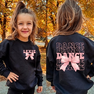 May include: Black sweatshirt with the word "DANCE" repeated three times in a retro font with a pink bow. The sweatshirt also has the name "Lexie" with a pink bow on the front.