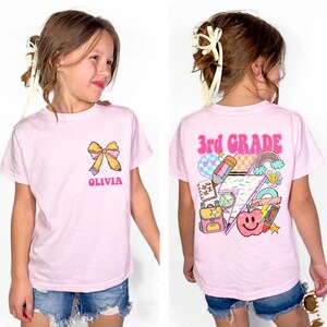 A young girl wearing a pink t-shirt with a cartoon design and the text "Third Grade" printed on it.