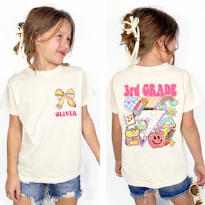 A young girl wearing a white t-shirt with a colorful graphic design and the text "Third Grade" printed on it.