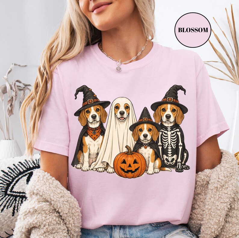 A woman wearing a pink t-shirt with a Halloween-themed graphic of four dogs dressed in costumes, including a ghost, skeleton, witch's hat, and pumpkin.