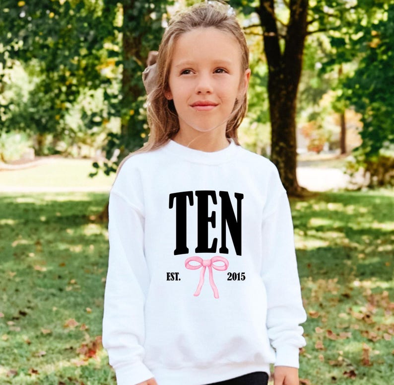 May include: A white long-sleeve sweatshirt featuring the black text TEN centered on the chest. Below the text is a small pink bow graphic flanked by the black text EST. on the left and 2015 on the right. The garment has a crew neckline.