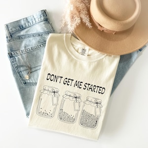 May include: Cream-colored t-shirt with the phrase "DON'T GET ME STARTED" and three jar illustrations. The shirt is paired with light-wash denim jeans, a tan hat, and a silver necklace. The shirt is made of a soft, comfortable material.
