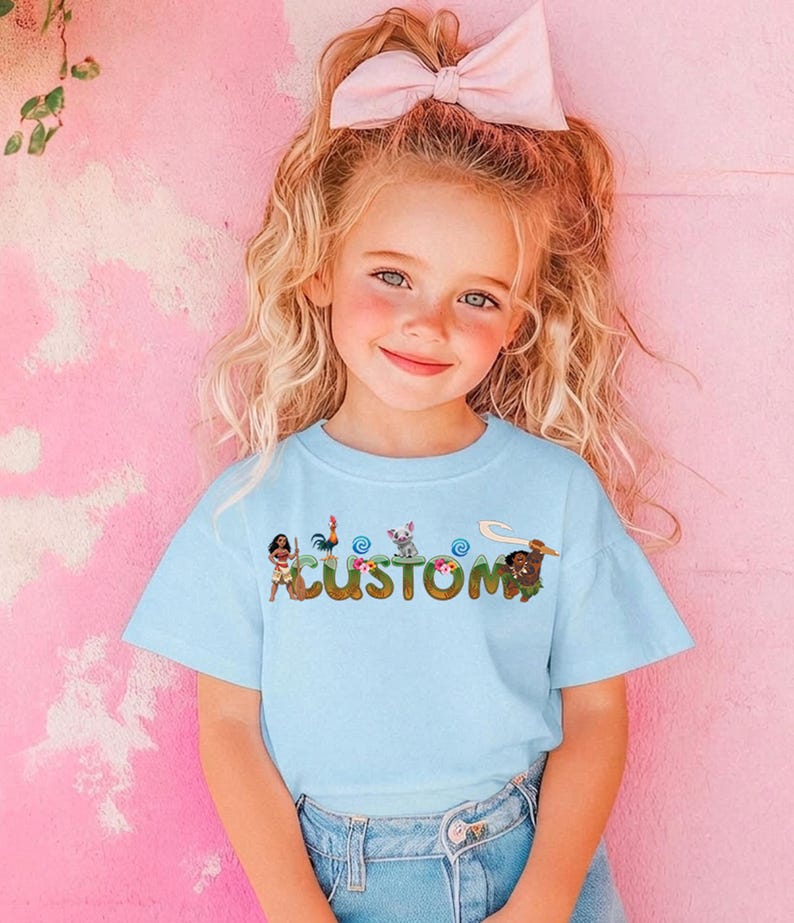A young girl with blonde hair wearing a light blue t-shirt with a cartoon character design and a pink bow in her hair.