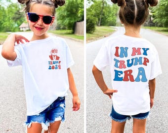 May include: A white t-shirt with a red, white and blue graphic of a person with the text "TRUMP 2024" on the front and the text "IN MY TRUMP ERA" on the back.