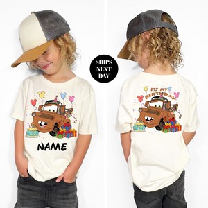 A child wearing a white t-shirt with a cartoon character from the Cars movie, specifically a tow truck, and the text "It's my birthday" printed on the back.
