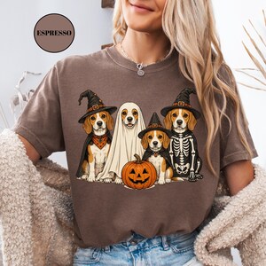 A woman wearing a t-shirt with a graphic design of four dogs dressed in Halloween costumes, including a ghost, skeleton, witch's hat, and pumpkin.