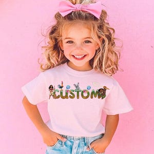A young girl with curly blonde hair is smiling and wearing a pink bow in her hair. She is wearing a white t-shirt with the word "CUSTOM" printed on it.