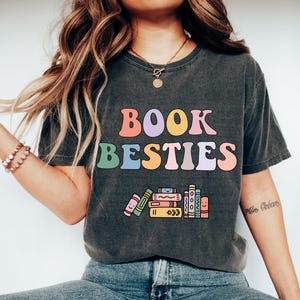 May include: A dark gray t-shirt with the text "BOOK BESTIES" in rainbow colors. The shirt features a stack of colorful books with smiling faces.